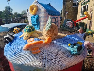 Beach-themed knitted postbox topper featuring cottage with blue roof, sea turtle, fish, badger, crab and seagull on wave-patterned knitwork with residential street in background.