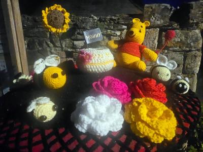 Winnie the Pooh knitted topper with bees, sunflower, honey pot and flowers on black postbox lid beside stone wall, with Hunny handwritten sign visible among the decorations.
