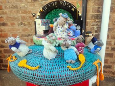 Knitted sheep scene titled Upton Rock showing seven mouse figures in sweaters playing musical intruments as a band around a central stage with green backdrop against brick wall, displayed on teal crocheted surface.