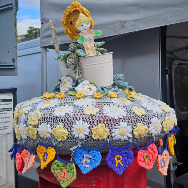 A red pillar postbox in Settle decorated with yarn bombing. On top sits a white crocheted flowerpot containing knitted figures of Bill and Ben the Flowerpot Men alongside Little Weed, the smiling weed character from the classic BBC children’s programme. The postbox lid is covered by a grey crochet doily with white and yellow daisies and green foliage. Around the rim hangs a garland of multicoloured crocheted hearts, each embroidered with a letter spelling out FLOWERPOT MEN.