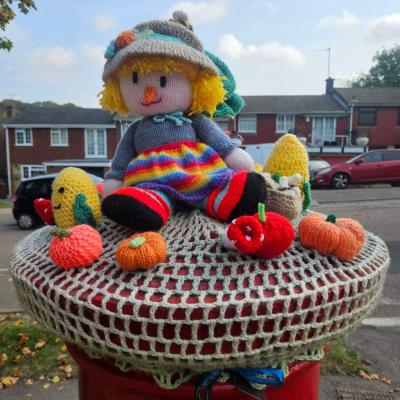 Knitted scarecrow doll in a pompom hat and rainbow skirt, surrounded by knitted corn, apples and pumpkins on a cream crochet base.