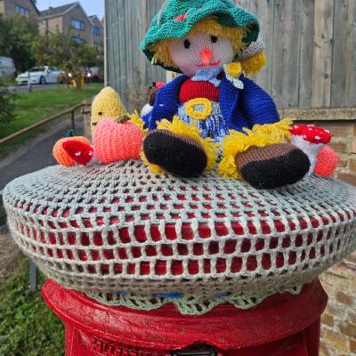 Knitted topper scarecrow sits in his green hat and blue jacket, with toy pumpkins and a mushroom beside him, edged with scalloped lace trim.