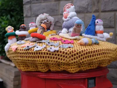 Knitted figures of people, fish, and sea creatures arranged on a crocheted yellow cover over a red post box, with a stone wall and greenery in the background.