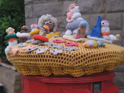 Knitted figures of people, fish, and sea creatures arranged on a crocheted yellow cover over a red post box, with a stone wall and greenery in the background.