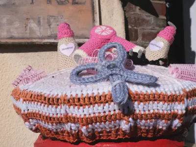 A crocheted postbox topper featuring hairdressing items including pink hair curlers, gray scissors, shampoo and conditioner bottles with labels, and pink hair rollers arranged on a white base with orange and white striped edging.