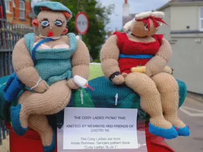 Two large knitted dolls dressed in swimsuits sitting on a red post box, one in turquoise and one in red, holding picnic items. A sign beneath them reads The Cosy Ladies Picnic Time, Knitted by members and friends of Eastry WI. The dolls are plump and cheerful, with detailed clothing and accessories, and are set against a street and building background.