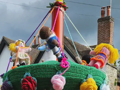 Colourful knitted figures of children dancing around a maypole on the top of a red post box. Each child holds a ribbon extending from the pole; the green base is decorated with large knitted flowers. The background shows rooftops and a cloudy sky.