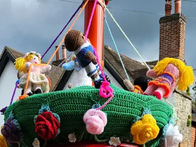 Colourful knitted figures of children dancing around a maypole on the top of a red post box. Each child holds a ribbon extending from the pole; the green base is decorated with large knitted flowers. The background shows rooftops and a cloudy sky.