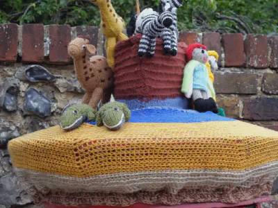 Knitted Noah's Ark scene on a post box. The decoration includes a knitted ark with black and white zebras, a chicken perched on top, two people in raincoats and boots, and a crocodile. The display sits in front of a brick wall and leafy trees.