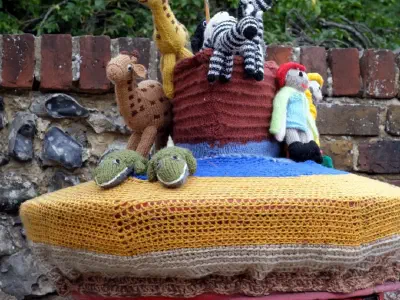 Knitted Noah's Ark scene on a post box. The decoration includes a knitted ark with black and white zebras, a chicken perched on top, two people in raincoats and boots, and a crocodile. The display sits in front of a brick wall and leafy trees.