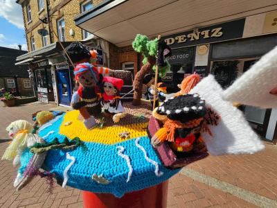 A large knitted pirate ship display with colorful yarn figures on a brick-paved street. The ship features a blue and yellow knitted hull with white foam-like waves, brown masts with rigging, and several knitted pirate characters wearing traditional hats and clothing. A large white knitted swan or seagull figure extends from the right side of the scene. The display sits on a pedestrian shopping street with traditional English storefronts visible in the background, including DENIZ shop signage.