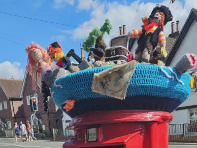 A pirate-themed knitted scene atop a red post box. The display features multiple pirates, a treasure chest, a shark, a palm tree, and a mermaid with pink hair, all crafted with yarn and arranged on a blue crocheted base, with houses and a traffic light in the background.