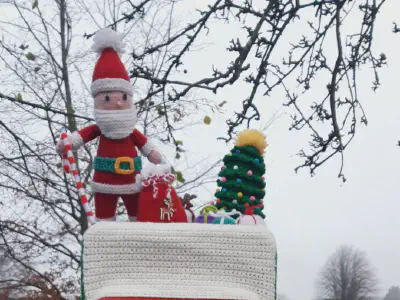 A red British postbox topped with a crocheted Santa Claus, Christmas tree, and presents, all on a crocheted snowy cover, set in a park with leafless trees and houses in the background.