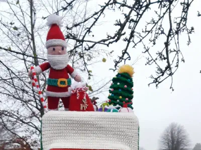 A red British postbox topped with a crocheted Santa Claus, Christmas tree, and presents, all on a crocheted snowy cover, set in a park with leafless trees and houses in the background.