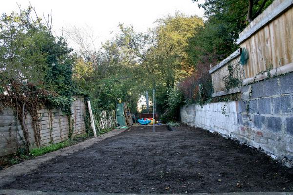 Freshly prepared garden bed with dark topsoil spread across narrow plot, ready for seeding, flanked by wooden fence and brick wall