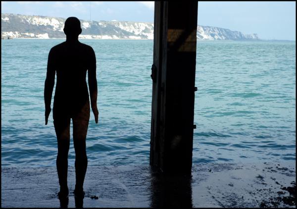 Silhouette of a person standing in shallow water looking out at turquoise sea, with white chalk cliffs visible in the distance under a clear sky. The figure is framed by what appears to be a dark architectural structure or pier support on the right side of the image.