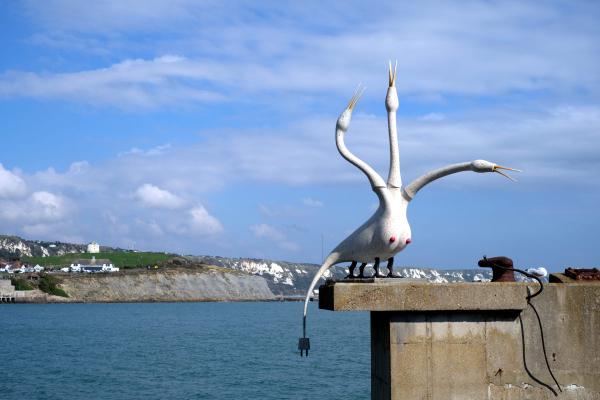 Sculptural art installation of a stylized white bird or crane-like figure positioned on a concrete ledge overlooking turquoise water. The sculpture has long, curved neck and limbs extended upward, with white chalk cliffs visible across the water in the background under blue skies with white clouds.