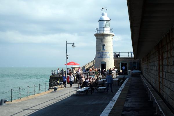 White lighthouse with cylindrical tower and lantern room sits at the end of a stone pier or breakwater. Visitors can be seen walking along the pier and gathered around the lighthouse base. The structure appears to be built from light-colored stone blocks, and the sea extends to the horizon under partly cloudy skies.