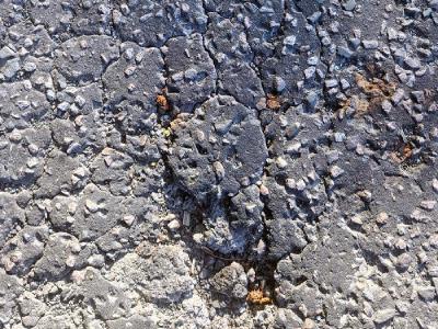Budgetary Contraints Close-up of deteriorated asphalt showing deep cracking with exposed aggregate and loose stones scattered across the broken surface.