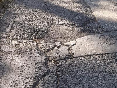 Quality Control Large pothole with surrounding radial cracking on a sunlit section of Labour in Vain Road, casting long shadows across the damaged pavement.