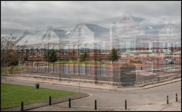 A ghostly overlaid photograph showing the ruins and archaeological remains of the former site of Invicta Works, with the transparent overlay of a Victorian red brick institutional building with distinctive chimneys and dormer windows. Modern residential housing is visible in the background, with construction work and excavation visible in the foreground area.