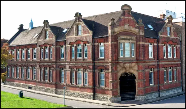A well-preserved Victorian red brick institutional building with distinctive architectural features including arched windows, ornate stonework, dormer windows with slate roofs, and a prominent arched entrance doorway. The building has two stories with a basement level visible, constructed in typical 19th-century style architecture.