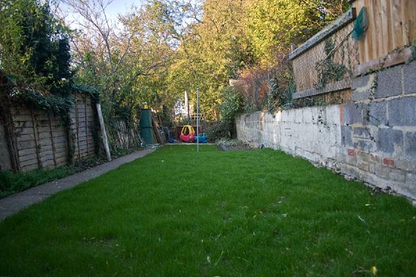 Established lawn in narrow garden with green grass, bordered by wooden fence on left and weathered brick wall on right, with play equipment visible at far end