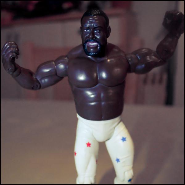 Action figure of professional wrestler Junkyard Dog wearing his signature white tights decorated with red and blue stars, shown in a flexing victory pose with arms raised and fists clenched, displaying his muscular physique and distinctive mohawk hairstyle