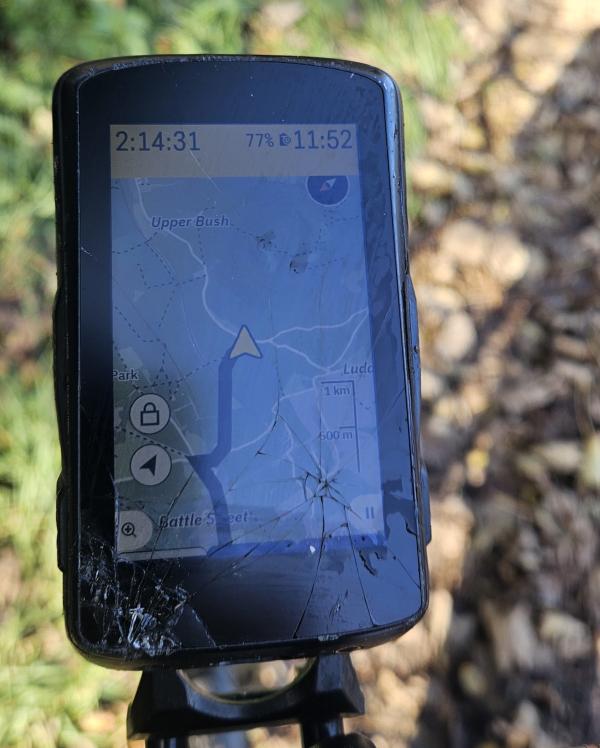 A Hammerhead Karoo 2 GPS cycling computer mounted on handlebars, showing a cracked screen with visible spider web fractures across the lower portion. The device is still operational and displays a map interface showing Upper Bush area, with battery at 77%, time reading 2:14:31, and navigation controls visible on the left side. The screen damage is concentrated in the bottom left corner with radiating cracks.