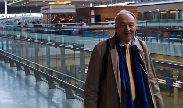 Ken Livingstone in business attire (suit jacket and tie) walking through St Pancras Station concourse, with glass railings, and contemporary architecture. He’s carrying an umbrella.