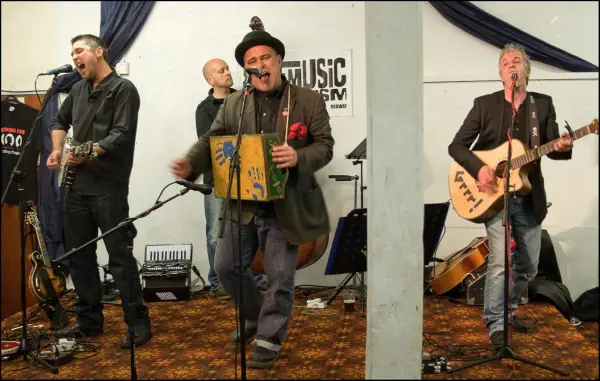 Four musicians performing live on stage at what appears to be a music venue. From left to right: a vocalist singing into a microphone while holding a guitar, a bald drummer in the background, a central performer wearing a black hat and jacket singing into a microphone while holding a colorful patchwork instrument or prop, and a guitarist on the right playing an acoustic guitar. The stage has white walls with black draping, a MUSIC sign visible in the background, and various musical equipment including amplifiers and microphones scattered across a patterned carpet floor.