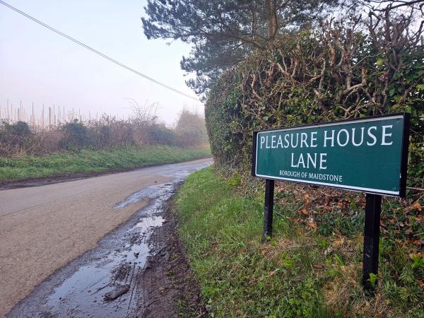 A green street sign reading “Pleasure House Lane, Borough of Maidstone” mounted on black posts beside a narrow, wet rural lane disappearing into misty farmland, with muddy verges and hedgerows on a damp winter morning