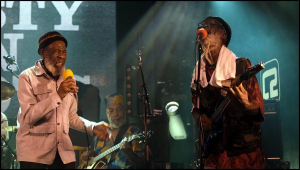 Two members of British reggae band Misty In Roots performing on stage during their 50th anniversary tour. The musician on the left holds percussion while wearing a pink button-up shirt and cap, while the guitarist on the right wears pink and performs with orange ear protection. Stage lighting creates a blue-green atmospheric backdrop.