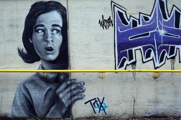 Monochrome street art of a surprised woman peeking from behind a wall, painted beside bold purple graffiti letters, with a bright yellow handrail running across the lower third of the image.