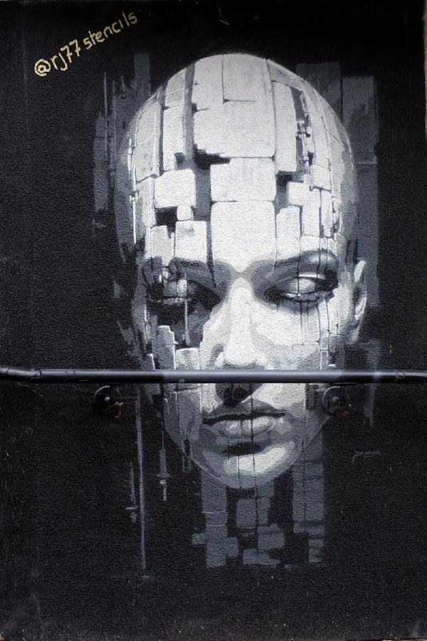 Black-and-white mural of a solemn human face on a dark wall, composed of pixel-like blocks, with a horizontal metal pipe crossing at mouth level and @rj77stencils stenciled in yellow at top left.