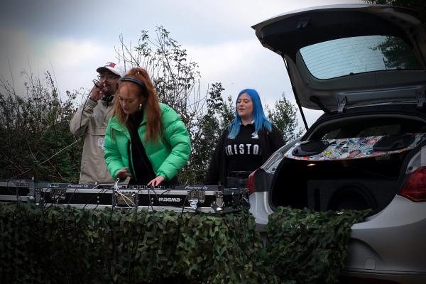 Outdoor DJ setup in front of a hatchback car draped in camouflage netting, featuring a female DJ in a green puffer jacket mixing on a Pioneer controller, flanked by two assistants against an overcast sky.
