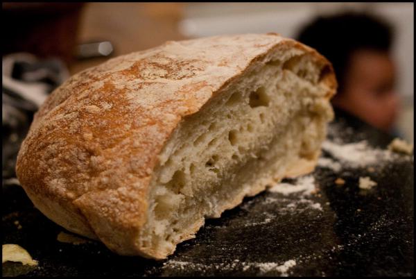 A close-up photograph of a rustic artisanal bread loaf with a golden-brown crust, cut to reveal its airy, holey interior crumb structure. The bread sits on a dark wooden cutting board with scattered flour, suggesting this is freshly baked no-knead bread.
