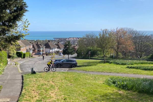 Two audax cyclists grinding up a steep suburban hill above the Kent coast, with houses and a bright blue sea visible in the distance.