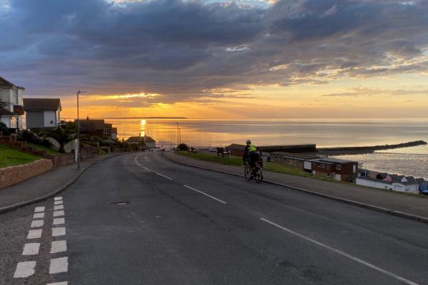 Audax rider on a coastal road out Herne Bay at sunset, with golden light reflecting across a calm sea beneath dramatic clouds.