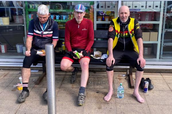 Three tired audax riders sitting outside a petrol station during a control stop, eating snacks and rehydrating, one barefoot with shoes off beside him.