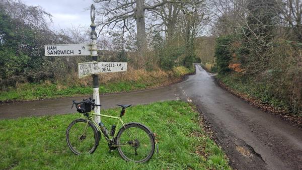 Touring bike leaned against a traditional fingerpost sign at a rural crossroads showing directions to Ham, Sandwich, Finglesham and Deal.