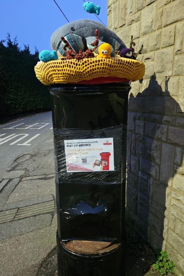 A black-wrapped postbox with a colourful knitted sea-life topper and a sign reading “Out of service – postbox of the future coming soon,” standing beside a stone wall at dusk.