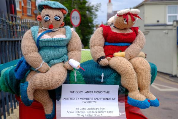 Two large knitted dolls dressed in swimsuits sitting on a red post box, one in turquoise and one in red, holding picnic items. A sign beneath them reads The Cosy Ladies Picnic Time, Knitted by members and friends of Eastry WI. The dolls are plump and cheerful, with detailed clothing and accessories, and are set against a street and building background.