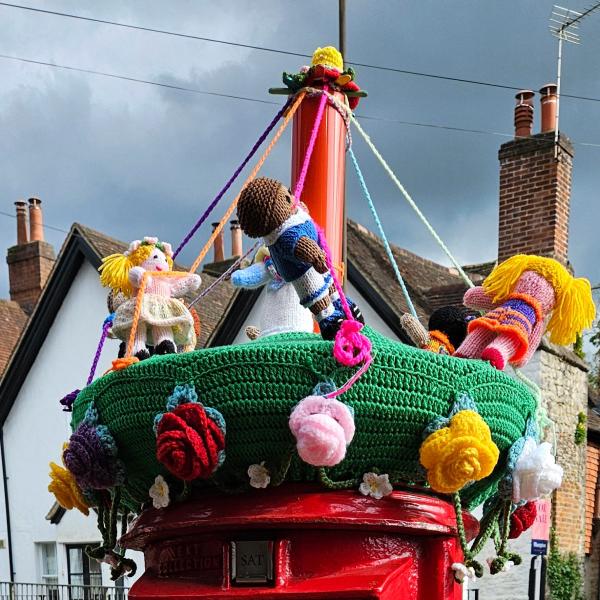Colourful knitted figures of children dancing around a maypole on the top of a red post box. Each child holds a ribbon extending from the pole; the green base is decorated with large knitted flowers. The background shows rooftops and a cloudy sky.