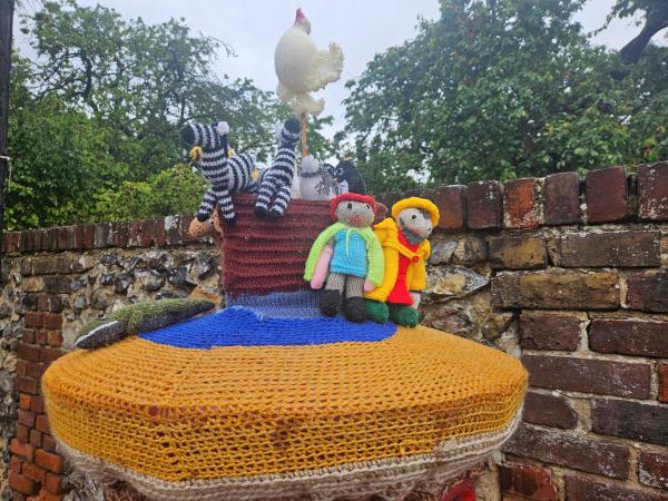 Knitted Noah’s Ark scene on a post box. The decoration includes a knitted ark with black and white zebras, a chicken perched on top, two people in raincoats and boots, and a crocodile. The display sits in front of a brick wall and leafy trees.