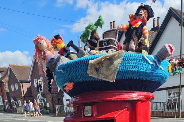 A pirate-themed knitted scene atop a red post box. The display features multiple pirates, a treasure chest, a shark, a palm tree, and a mermaid with pink hair, all crafted with yarn and arranged on a blue crocheted base, with houses and a traffic light in the background.