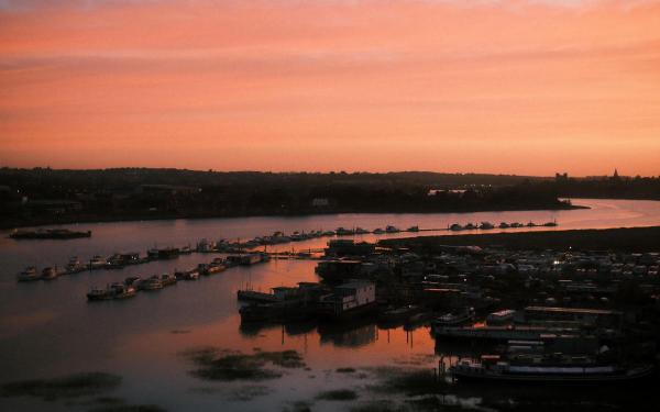 Boats moored along a tidal river at dawn, their windows lit, with a pink-and-gold sky reflected across the calm water and the silhouettes of towers on the far shore.