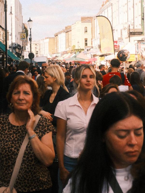 A dense crowd of shoppers filling Portobello Road on a sunny day, with market stalls, lamp posts and tall pastel-coloured buildings behind.