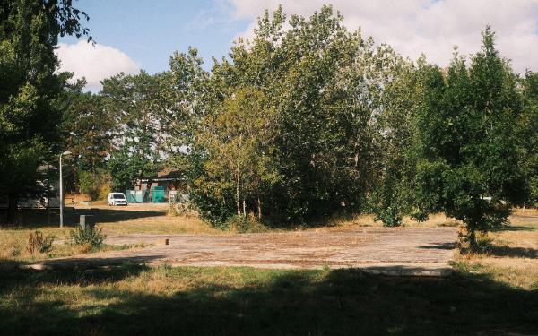 A bare concrete slab surrounded by rough grass and mature trees, with a caravan and a small portable building visible behind it.