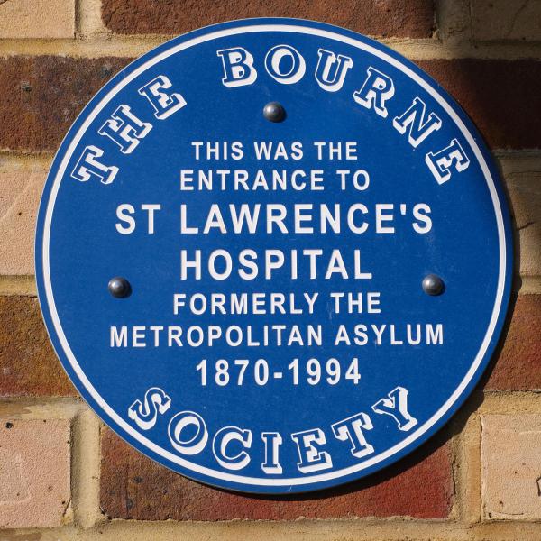 Round blue heritage plaque mounted on a brick wall, reading: The Bourne Society - this was the entrance to St Lawrence’s Hospital, formerly the Metropolitan Asylum, 1870-1994.